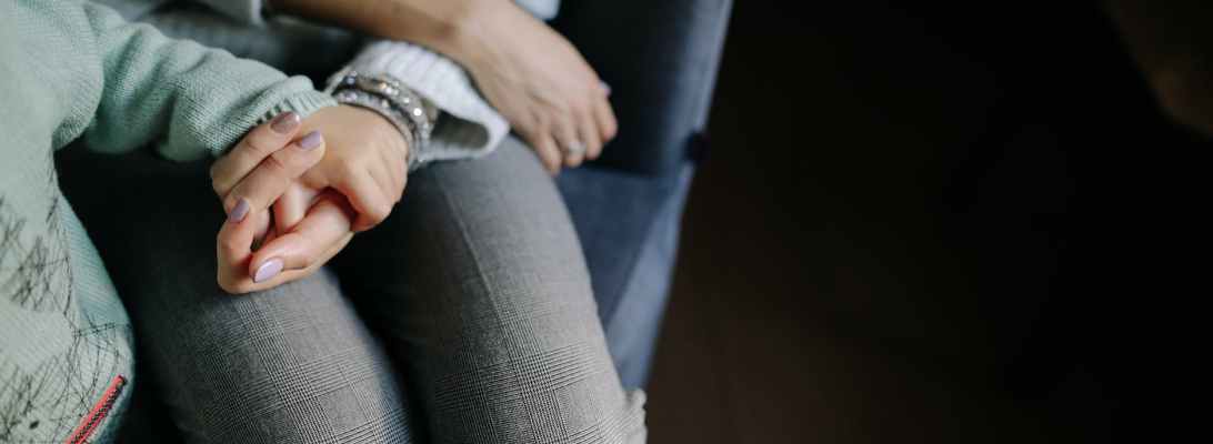 Woman sitting in a grey long sleeved shirt holding mother's hand in blue long sleeved shirt