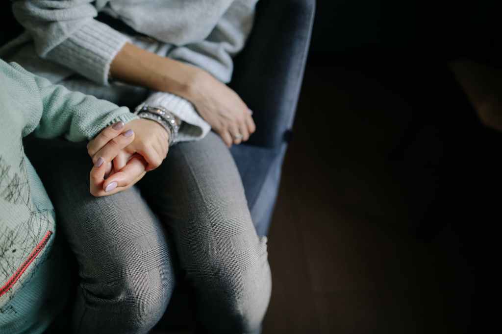 Woman sitting in a grey long sleeved shirt holding mother's hand in blue long sleeved shirt