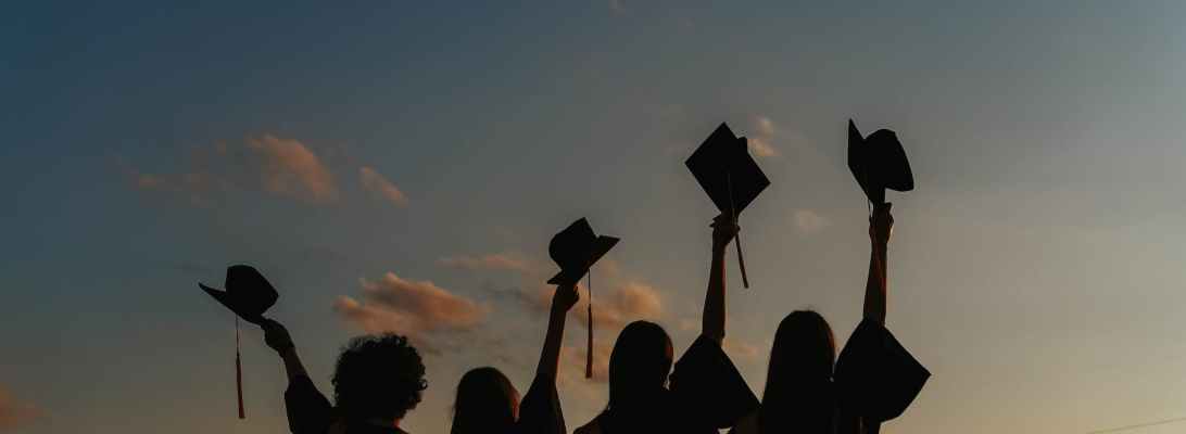 Silhouette of four students raising their graduation hats with a sunset behind them