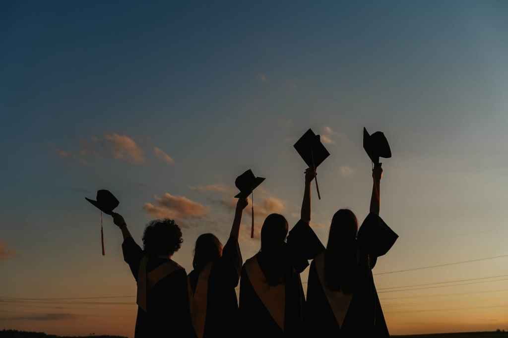Silhouette of four students raising their graduation hats with a sunset behind them