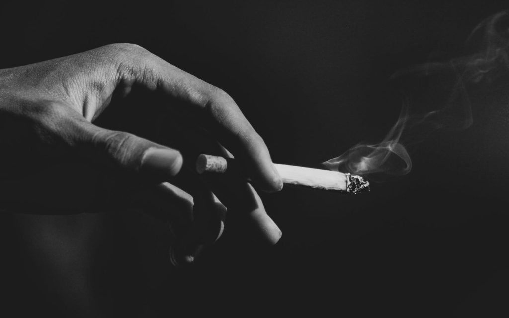 A black and white photo of a hand holding a lit cigarette, with smoke rising from the end.