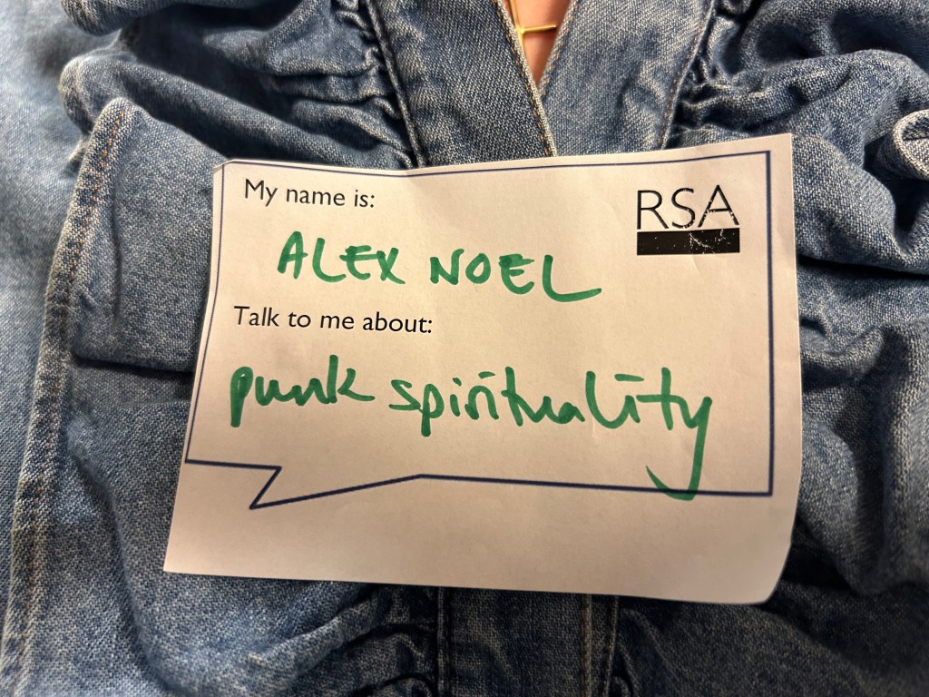 A sticker on a shirt saying: My name is Alex Noel, talk to me about Punk Spirituality. With the RSA (Royal Society of Arts) logo. 